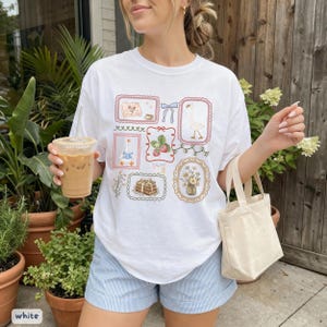 May include: White t-shirt featuring a collection of framed illustrations with a goose, strawberries, and flowers. The shirt has a relaxed fit and is paired with light blue striped shorts. A beige tote bag and iced coffee are also visible.