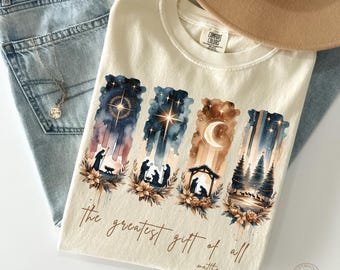 May include: Ivory-colored t-shirt with a watercolor design featuring Nativity scenes and the text "the greatest gift of all" and "matthew 1:21". The shirt has a crew neck and is made of a soft material. The shirt is folded on top of blue jeans.