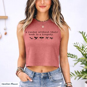 May include: Heather mauve crop top with the text "a reader without their book is a tragedy." and black graphic illustrations. The top is sleeveless and made of a soft material. The model is wearing blue jeans.