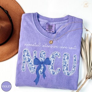 May include: A violet Comfort Colors t-shirt with the text "neonatal intensive care unit" and "NICU" in a floral design. A blue bow is centered below the letters. A gold necklace and a brown hat are in the background.