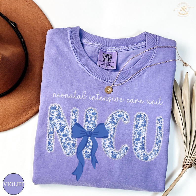 May include: A violet Comfort Colors t-shirt with the text "neonatal intensive care unit" and "NICU" in a floral design. A blue bow is centered below the letters. A gold necklace and a brown hat are in the background.