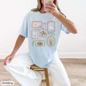 May include: Light blue t-shirt featuring a collection of framed illustrations. The illustrations include a goose, books, strawberries, and flowers. The shirt has a crew neck and short sleeves. The word "chambray" is printed at the bottom left.
