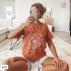 May include: Burnt orange t-shirt featuring a floral design and the text "IT IS WELL with my soul." The graphic includes flowers and a butterfly. The shirt is worn with white shorts and socks.