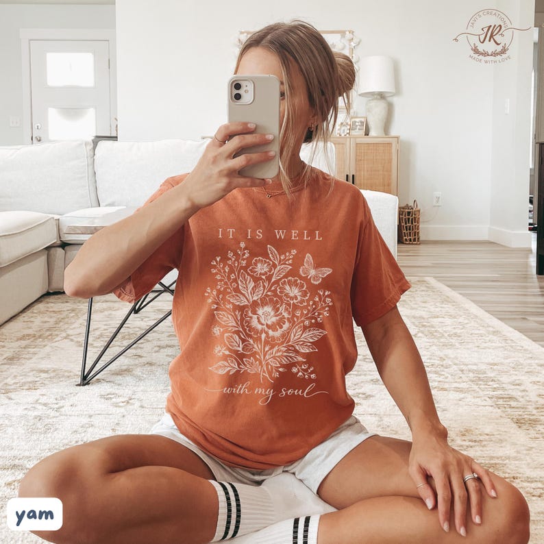 May include: Burnt orange t-shirt featuring a floral design and the text "IT IS WELL with my soul." The graphic includes flowers and a butterfly. The shirt is worn with white shorts and socks.