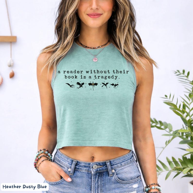 May include: Heather dusty blue crop top with the text "a reader without their book is a tragedy." and black graphic illustrations. The top is sleeveless and made of a soft material.