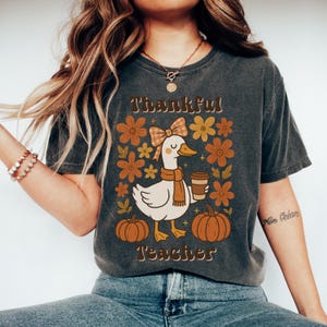 May include: Dark gray t-shirt with a fall-themed graphic. The design features a white duck wearing a scarf and holding a coffee cup, surrounded by flowers and pumpkins. Text reads "Thankful Teacher".