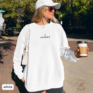 May include: White sweatshirt with the word "YAHWEH" printed in black, with a cross above the text. The sweatshirt is worn with a white cap and sunglasses. A white purse is over the shoulder. A coffee cup is held in the hand.