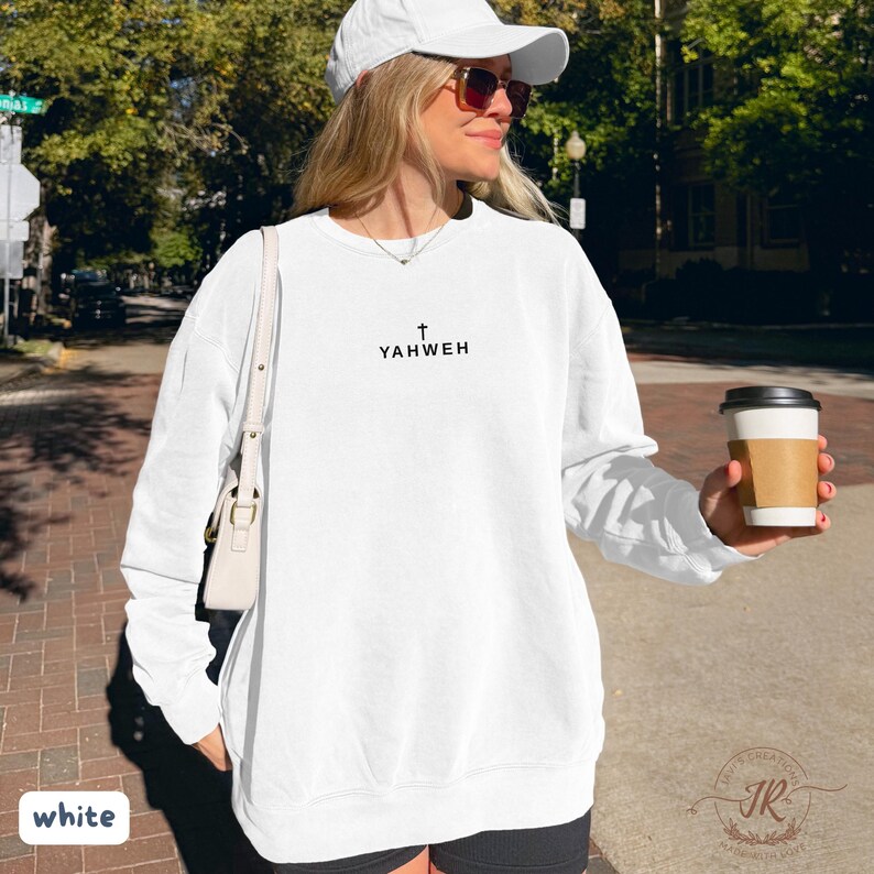 May include: White sweatshirt with the word "YAHWEH" printed in black, with a cross above the text. The sweatshirt is worn with a white cap and sunglasses. A white purse is over the shoulder. A coffee cup is held in the hand.