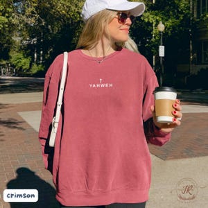 May include: Crimson-colored sweatshirt with the word "YAHWEH" and a cross printed in white. The sweatshirt is a crew neck style and has a relaxed fit. The image also shows a white purse strap and a coffee cup.