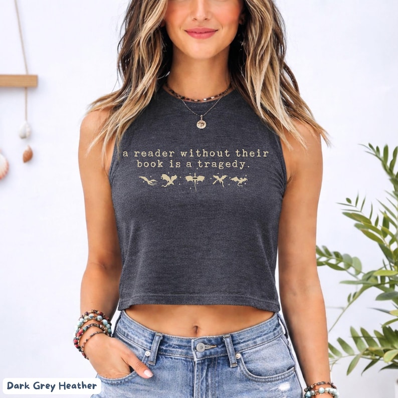 May include: Dark grey heather crop top with the quote "a reader without their book is a tragedy" in gold lettering. The top features a sleeveless design and a crew neck. The text is accompanied by small illustrations. The model is wearing blue jeans.