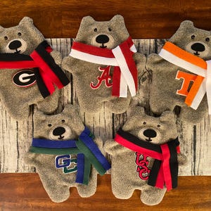 May include: Five gray plush teddy bears wearing different colored scarves with college logos. The logos are for the University of Georgia, Alabama, Tennessee, Clemson, and USC.