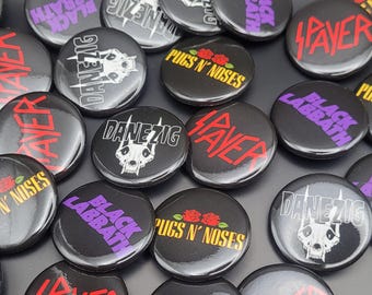 Op de afbeelding: Een verzameling ronde zwart-witte knoppen met verschillende bandlogo's, waaronder Slayer, Danzig, Black Sabbath en Pugs N' Noses. De knoppen zijn ongeveer 5 cm in diameter.