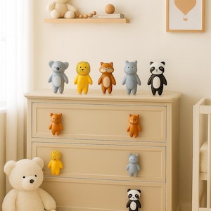 Può includere: Una cassettiera beige con pomelli a forma di animali e statuette decorative. Le statuette includono un koala, un leone, una volpe, un ippopotamo e un panda. Un grande orsacchiotto è sul pavimento e un'immagine incorniciata è appesa al muro.