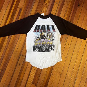 May include: A white and black baseball tee shirt with a graphic of a rat in front of a concert stage. The text "RATT" is printed in large letters above the graphic.