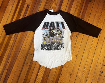 Puede incluir: Una camiseta de béisbol blanca y negra con un gráfico de una rata frente a un escenario de concierto. El texto "RATT" está impreso en letras grandes sobre el gráfico.