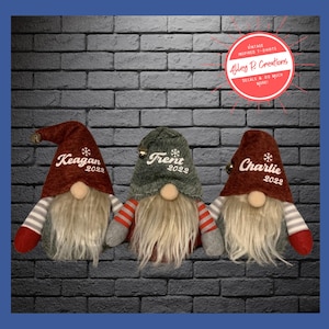 May include: Three personalized Christmas gnomes with red and gray hats. The gnomes are standing in front of a brick wall. The hats are embroidered with the names Keagan, Frent, and Charlie, and the year 2022.