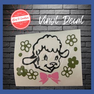 May include: A black and white vinyl decal of a cartoon lamb with a pink bow. The lamb is surrounded by green flowers.