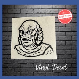 May include: A black and white vinyl decal featuring a stylized illustration of the Creature from the Black Lagoon. The image shows the creature's face with detailed lines and shading. The text "Vinyl Decal" is at the bottom.