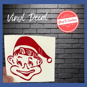 May include: Red vinyl decal of a smiling elf wearing a Santa hat. The decal is on a white background. Vinyl Decal. VINTAGE INSPIRED T-SHIRTS Alley R. Creations DECALS & SO MUCH MORE!