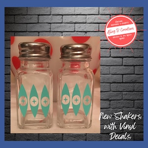 May include: Two clear glass salt and pepper shakers with teal and white vinyl decals. The shakers have silver lids. The text "New Shakers with Vinyl Decals" is visible below the shakers.