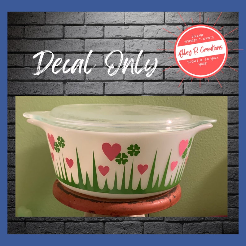 A white casserole dish with a green and pink floral design. The design features green grass, pink hearts, and green four-leaf clovers. The design is inspired by the vintage Pyrex Lucky in Love pattern. The dish is on a red stand. The text 'Decal Only' is written in white cursive above the dish. There is an accompanying logo for Alley R Creations in the upper right hand corner.