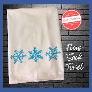 May include: White flour sack towel with three blue snowflake designs. The text "Flour Sack Towel" is printed in black below the snowflakes.