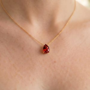 Peut inclure: Un collier en or avec un pendentif en forme de goutte d'eau, une pierre précieuse rouge foncé. Le pendentif est serti dans une lunette en or et est suspendu à une fine chaîne en or. Le collier est présenté sur une peau et un haut noir.