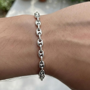 Könnte beinhalten: Ein silberfarbenes Armband mit einer Reihe von ovalen Gliedern, die durch kleinere, kreisförmige Glieder verbunden sind. Das Armband wird an einem Arm gezeigt, um sein Design hervorzuheben. Der Verschluss hat ein dekoratives Blumenmuster. Länge ca. 18 cm.