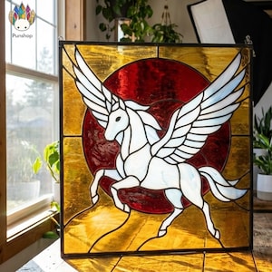 Op de afbeelding: Gebrandschilderd glaspaneel met een witte Pegasus met uitgespreide vleugels, tegen een rode cirkelvormige achtergrond. Het paneel is omlijst met goud en amberkleurig glas. Een decoratief item voor in huis, ongeveer 30 cm x 30 cm.