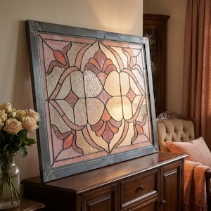 May include: A large, rectangular stained glass panel with a gray metal frame. The design features a symmetrical floral pattern in shades of pink, peach, and cream. The glass has a textured appearance, adding depth to the artwork. The panel is displayed on a dark wooden cabinet.