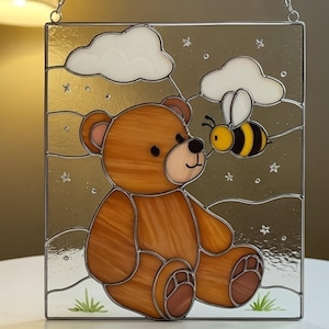 May include: A square stained glass panel depicting a brown teddy bear and a yellow and black bee. The bear is seated, with the bee hovering close by. White clouds and stars are in the background. The panel has a silver border.