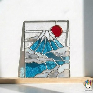 Peut inclure: Panneau en vitrail représentant le Mont Fuji avec un soleil rouge, une montagne bleue et des nuages blancs. L'œuvre est encadrée d'une bordure en métal argenté et repose sur une surface en bois. Le design est inspiré de l'art japonais.