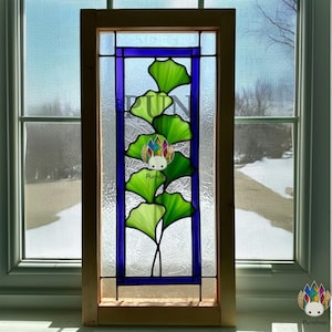 May include: Stained glass window panel featuring a ginkgo leaf design in shades of green and yellow. The leaves are set against a blue background within a rectangular wooden frame. The window is positioned in front of a window with a view of a snowy landscape.
