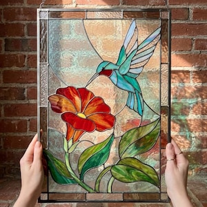 Pode incluir: Um painel retangular de vitral apresentando um beija-flor com asas azul-petróleo e brancas e garganta vermelha, pairando sobre uma grande flor vermelha com centro amarelo. O design inclui folhas verdes e é emoldurado em metal escuro.