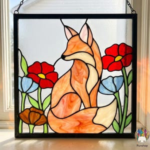 Pode incluir: Painel de vitral com uma raposa no meio de flores vermelhas e azuis. A raposa tem pelo laranja e creme, e as flores têm caules e folhas verdes. O painel é emoldurado em preto e pendurado por uma corrente.
