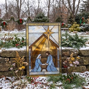 May include: A rectangular stained glass panel depicting a nativity scene with two figures in blue robes kneeling beside a manger under a large golden star. The panel features a warm amber background within a dark frame, showcasing intricate glass work.