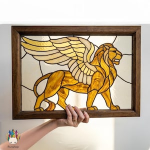 Op de afbeelding: Gebrandschilderd kunstwerk met een gouden gevleugelde leeuw in een donkere houten lijst. De leeuw heeft gedetailleerde vleugels en een koninklijke pose. Het glas gebruikt tinten goud, amber en crème. Het kunstwerk wordt door een persoon vastgehouden.
