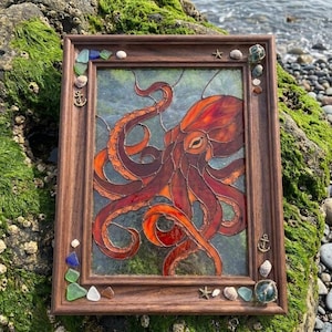 May include: A rectangular stained glass panel featuring a red and orange octopus, encased in a dark brown wooden frame. The frame is decorated with small sea-themed accents including seashells, sea glass pieces, and metal anchor charms.