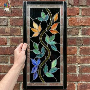 May include: Stained glass panel with a botanical design. It features leaves in blue, green, orange, and yellow against a black backdrop. The panel is framed in black with a decorative border. The panel is likely made of glass.