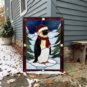 Pode incluir: Um painel de vitral retangular apresentando um pinguim usando um gorro de Pai Natal vermelho e branco e um cachecol vermelho. O pinguim está sobre uma superfície branca nevada, rodeado por pinheiros verdes contra um céu azul escuro.