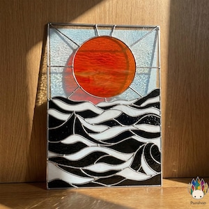 May include: Stained glass panel showing a sunset over the sea. The sun is a bright orange, and the waves are black and white. The panel is framed with a silver metal border.