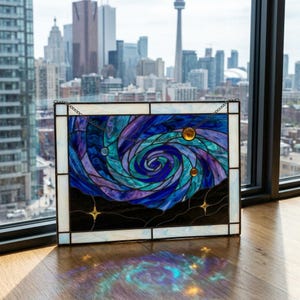 May include: A rectangular stained glass panel featuring a swirling galaxy design in shades of deep blue, purple, and teal. The piece includes small amber-colored circular accents and star shapes, framed by a white border with a metal hanging chain.