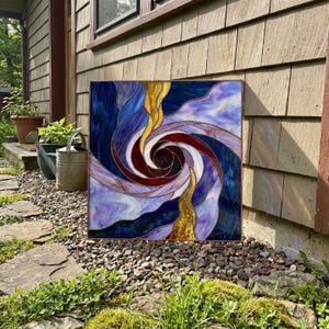 以下が含まれることがあります： 渦巻き状の模様が特徴の正方形のステンドグラスパネル。深い青、明るい紫、黄金色のガラス片が暗い色の金属フレームで繋ぎ合わされており、中央の暗い赤色の核に向かって回転するダイナミックな螺旋模様が描かれています。