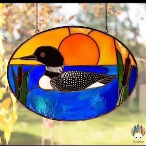 May include: Oval stained glass panel featuring a loon on a blue lake with a sunset background. The loon is black and white with a red eye. The sunset is orange and yellow. Cattails are on the sides. The panel is hanging by a chain.