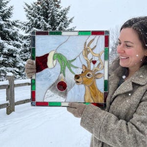 May include: A square stained glass panel featuring a green hand in a red sleeve holding a red ornament near a brown reindeer with a small red heart on its antlers. The panel is framed by a border of alternating red, white, and green glass pieces.