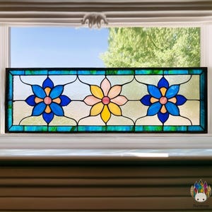 Könnte beinhalten: Rechteckige Glasfensterscheibe mit drei stilisierten Blumen. Die Blumen sind in Blau, Rosa und Gelb gehalten, mit schwarzen Umrissen. Das Panel hat einen blau-grünen Rand und zeigt einen Blick auf den Himmel.