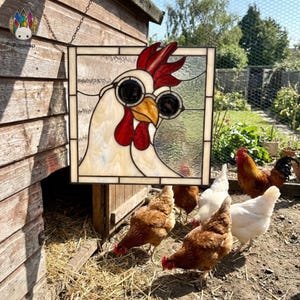 Könnte beinhalten: Ein Buntglasfenster mit einem Huhn, das eine schwarze Sonnenbrille trägt. Das Huhn hat einen roten Kamm und Kehllappen. Mehrere Hühner sind im Vordergrund, mit einem hölzernen Hühnerstall im Hintergrund. Das Fenster hängt an einer Kette.