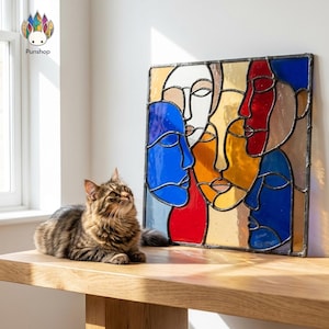 Op de afbeelding: Een kleurrijk glas-in-loodpaneel met abstracte gezichten in blauw, rood, geel en wit. Het kunstwerk is zwart omlijst en staat op een houten oppervlak. Er is ook een tabby kat aanwezig.