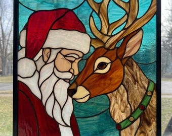 Puede incluir: Un panel de vidriera que muestra el perfil de Santa Claus con gorro rojo y barba blanca frente a un reno marrón con grandes astas. El fondo es de vidrio azul verdoso texturizado y la pieza está enmarcada con un borde de metal negro.