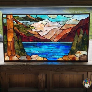 May include: Stained glass panel showing a landscape with a lake, mountains, and trees. The artwork uses blue, orange, green, and brown colours, and is framed in a black border. The panel is displayed on a wooden surface.